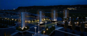Vista nocturna aérea de ingreso a complejo moderno con pilares luminosos verticales, espejos de agua y jardines, diseño Lineasur Arquitectos Cipolletti