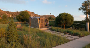 esidencia compacta sustentable al atardecer – Proyecto residencial Cipolletti, Río Negro. Casa con piedra local, paneles solares, turbina eólica, deck con fogón y jardín nativo integrado al paisaje de mesetas